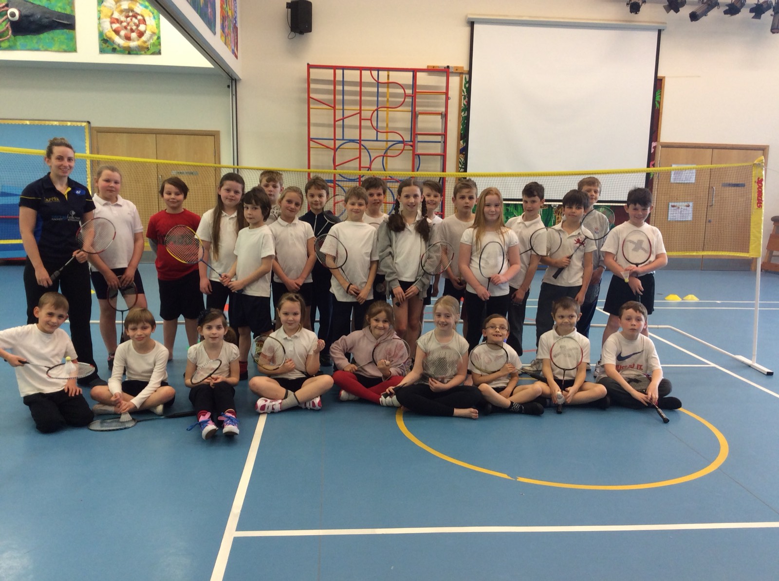 A primary school class holding badminton rackets on court with coach Kirsty Wilcox